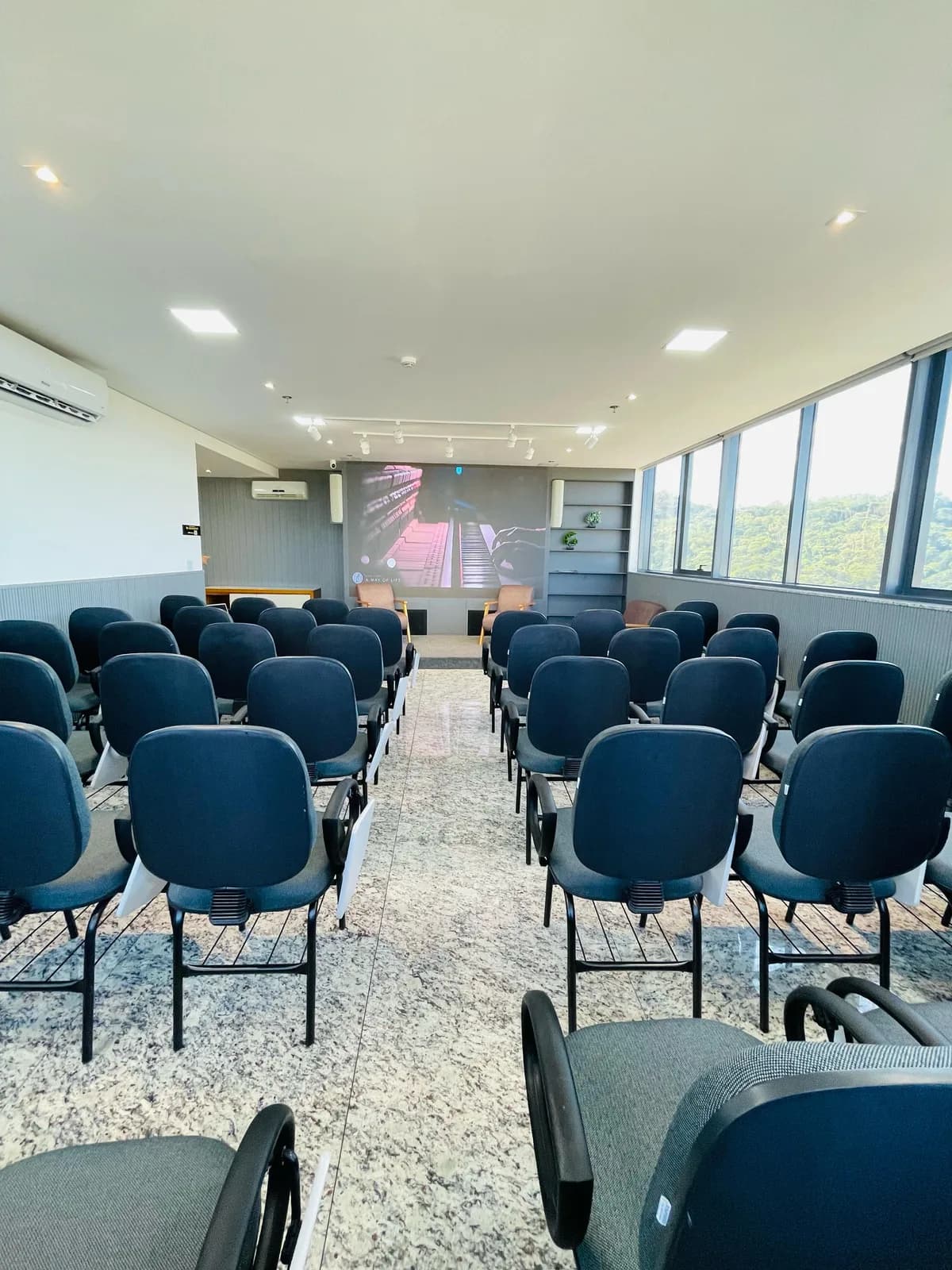 Auditório do Ipatinga Tower: fileiras de cadeiras de frente para o telão, com janelas amplas para a mata